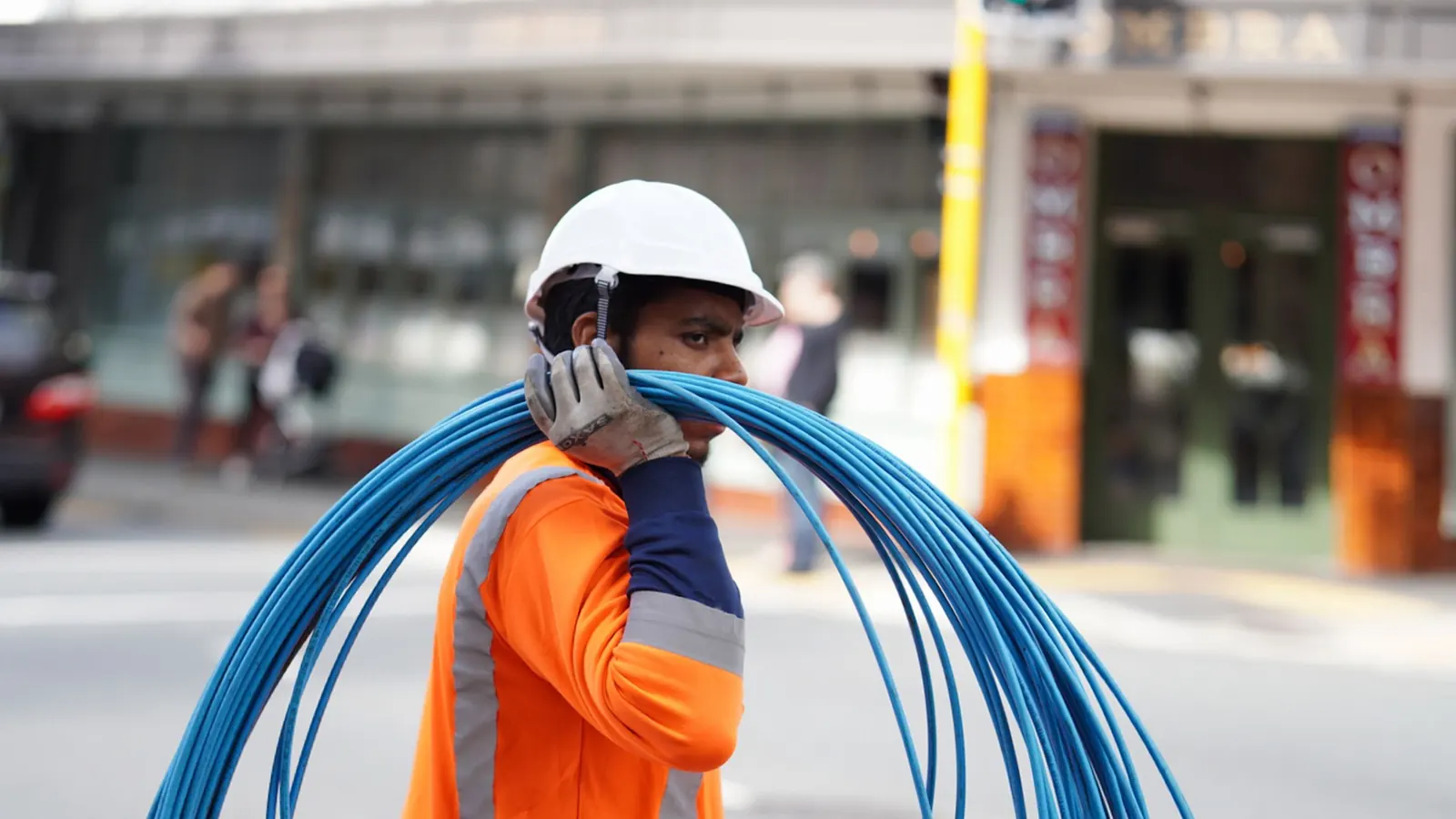 Tradesperson in hi-vis workwear carrying coiled blue cabling on a city street