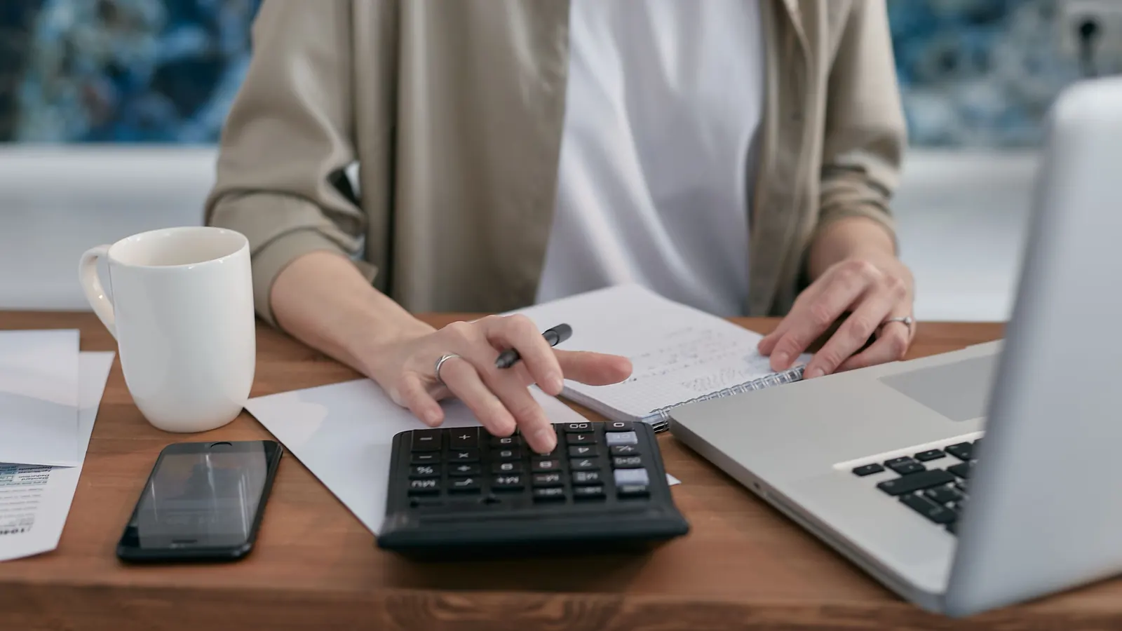 Hands typing on a desk calculator beside a laptop, a phone and a mug on a wooden desk