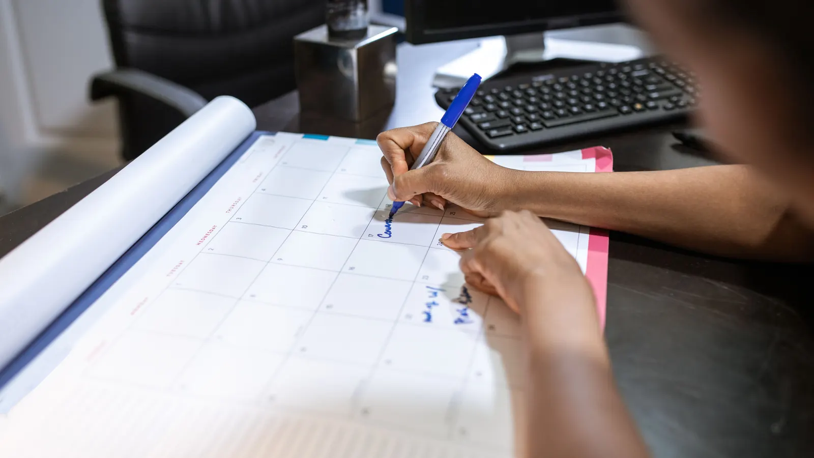 A hand marking dates on a large desk calendar with a blue pen
