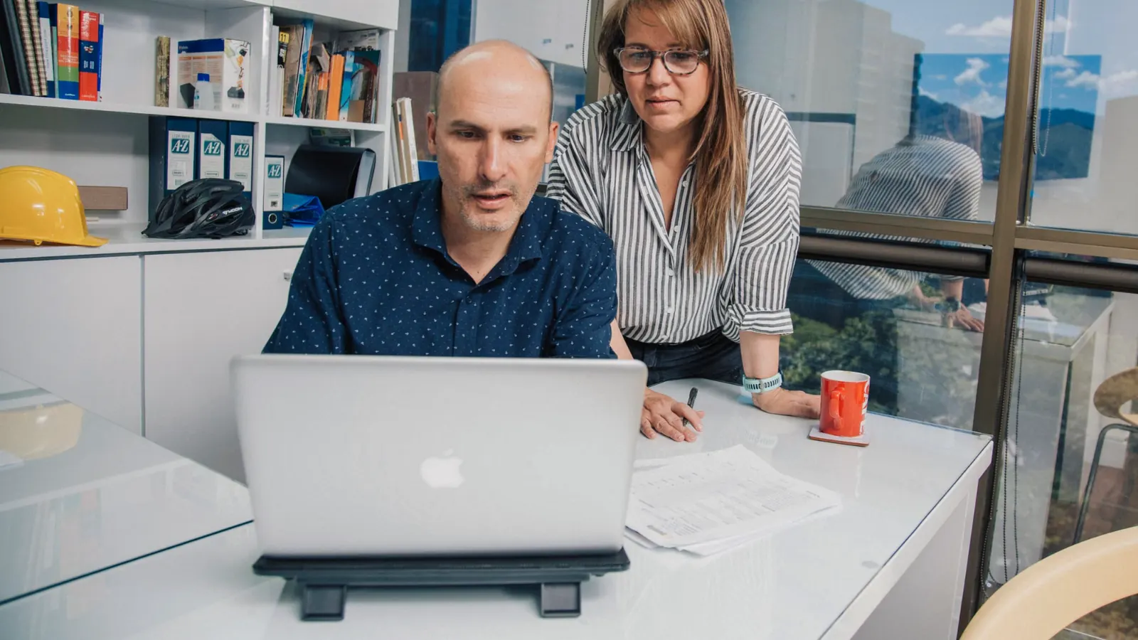 Two colleagues reviewing a laptop together in an office, with a hard hat visible on the shelf