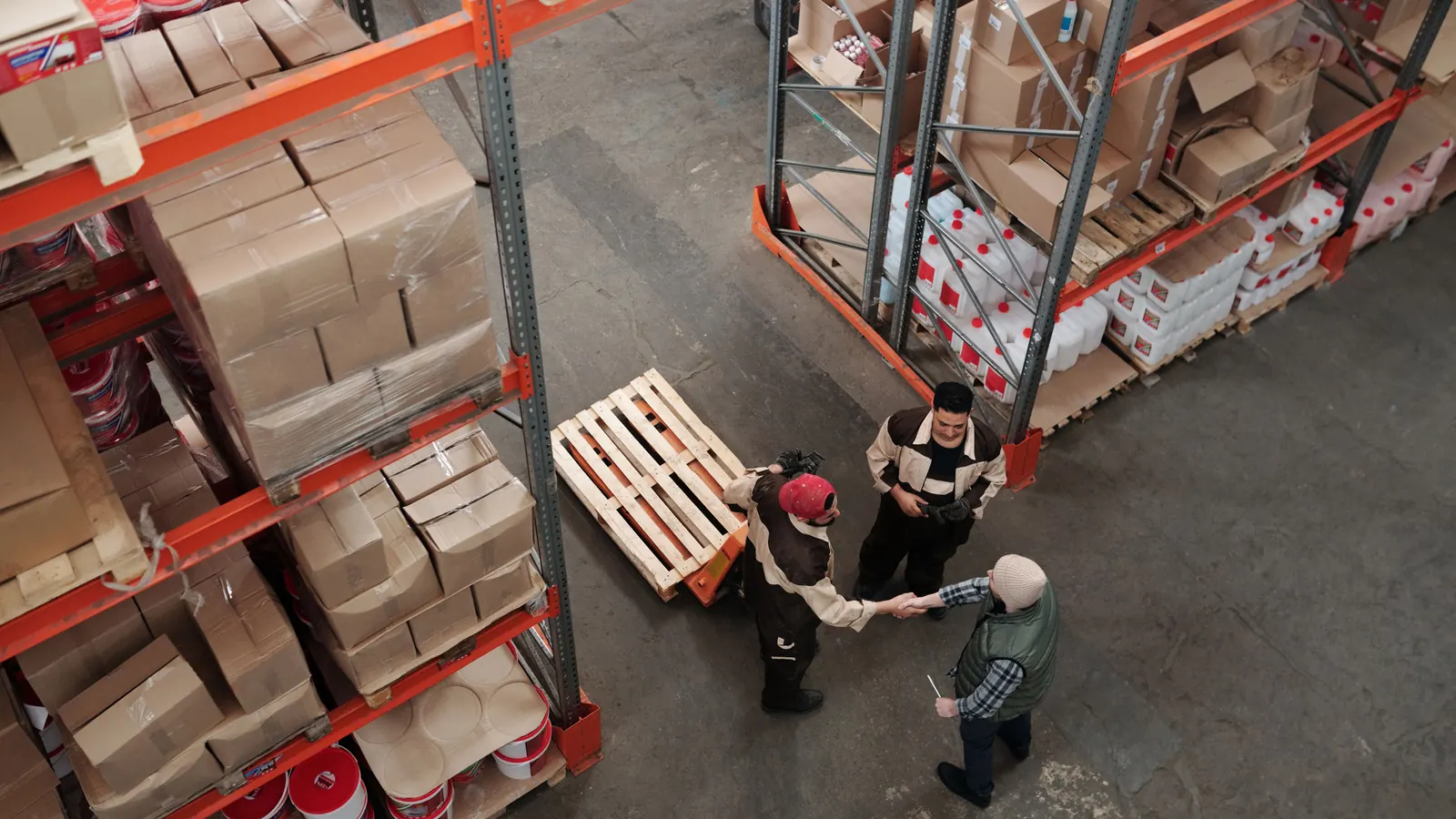 Two warehouse workers shaking hands beside stacked pallets of stock