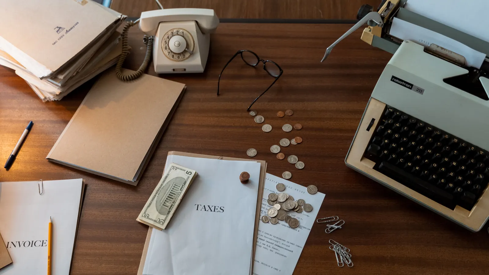 An overhead view of a desk with paperwork labelled Invoice and Taxes, loose coins, reading glasses and a typewriter
