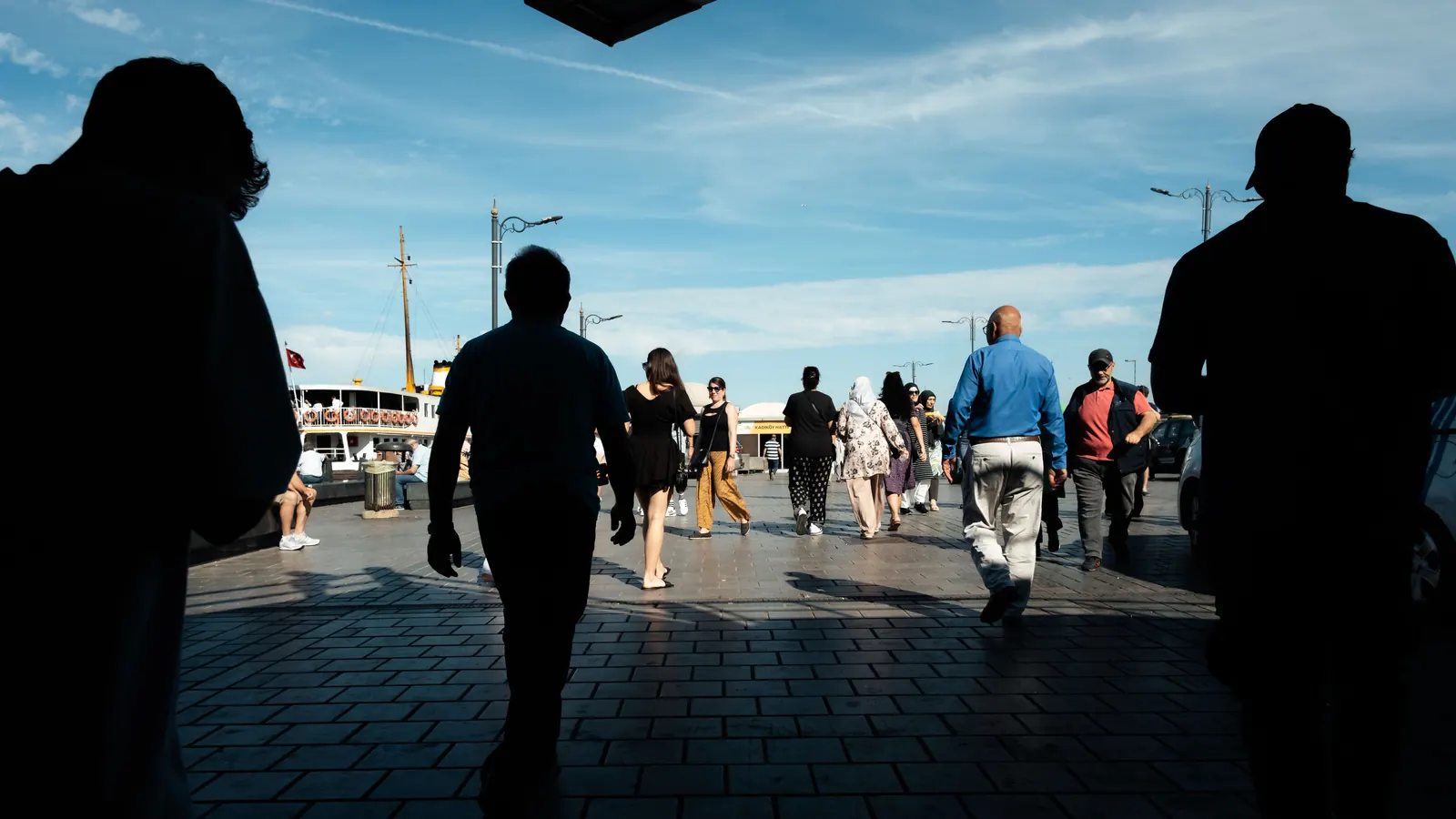 Silhouetted figures walking across a busy waterfront square in the morning sun