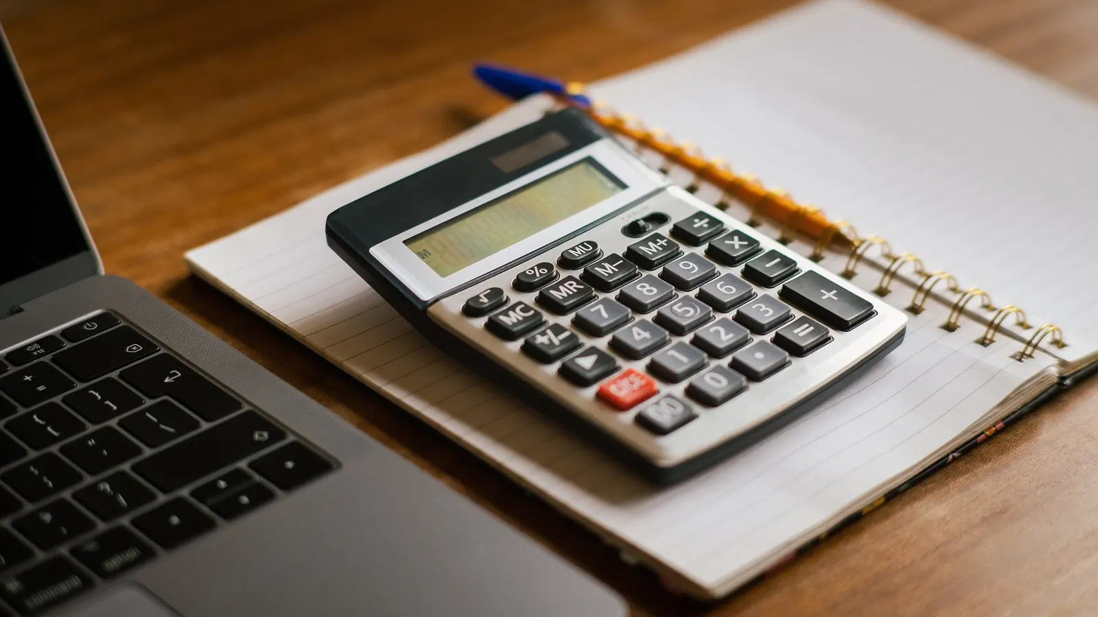 A calculator resting on an open notebook beside a laptop on a wooden desk