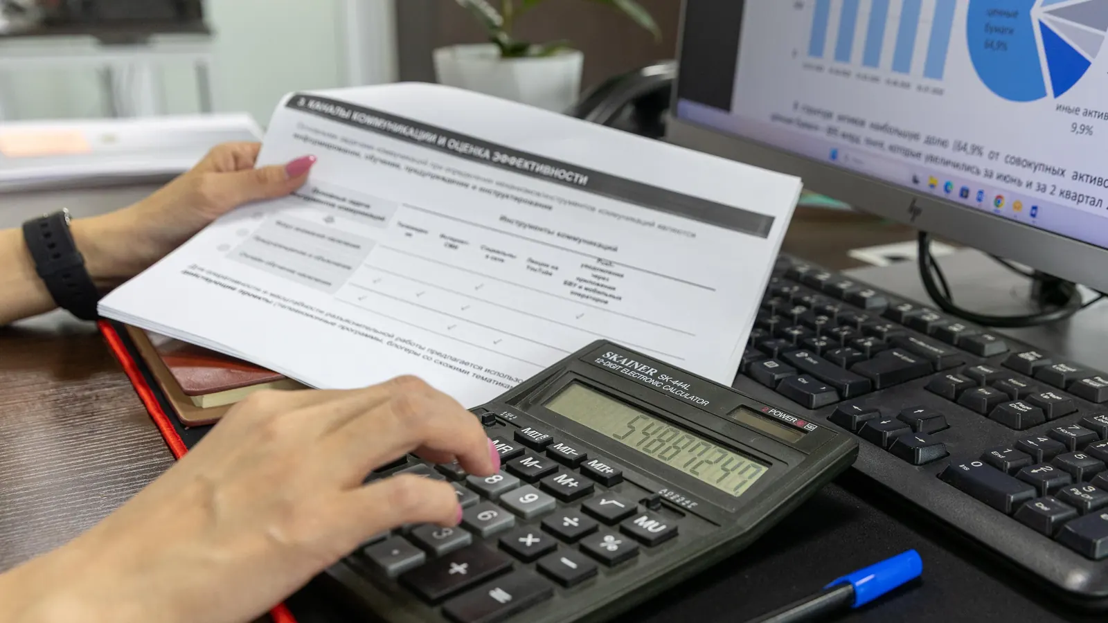 Hands entering figures on a desk calculator while holding a printed form beside a keyboard