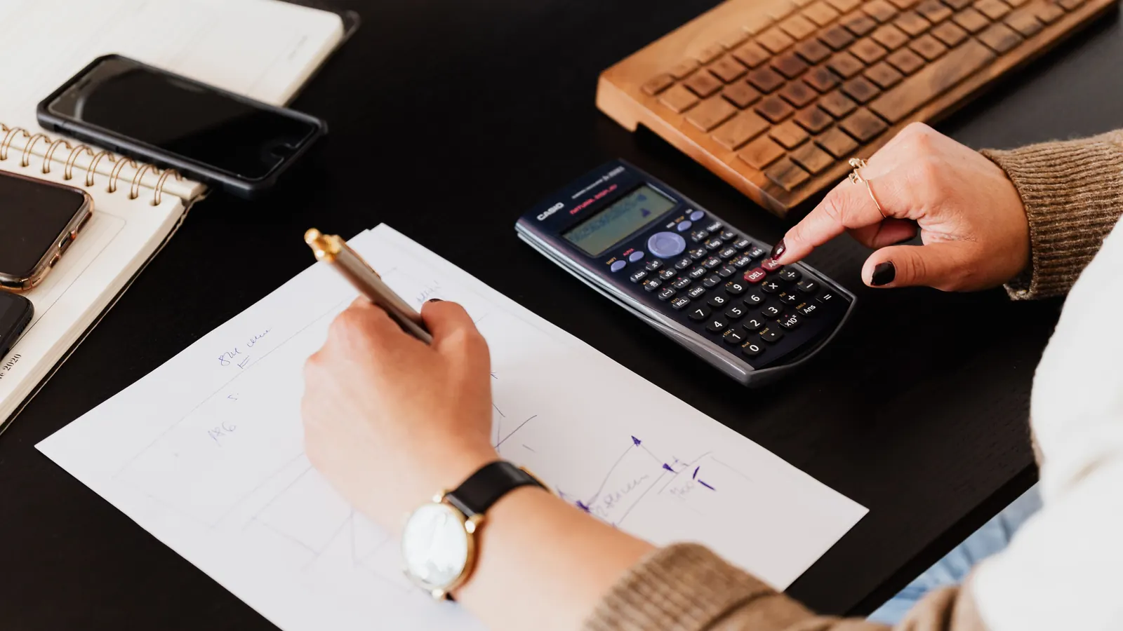 A person writing figures on paper with a pen while using a calculator at a dark desk