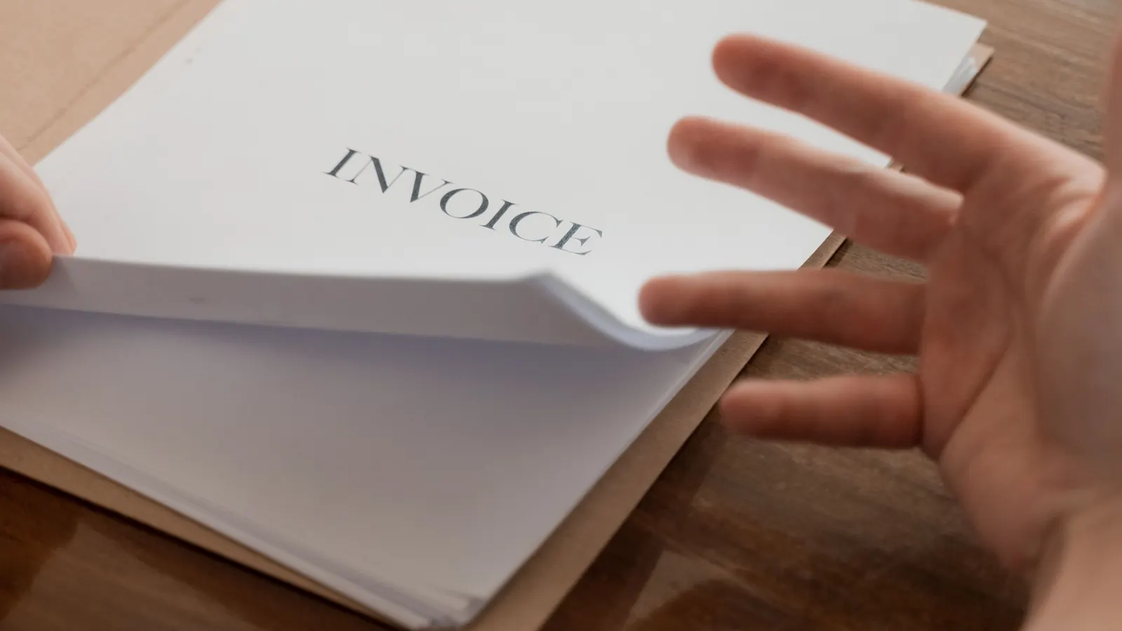 A hand leafing through a stack of printed invoices on a wooden desk