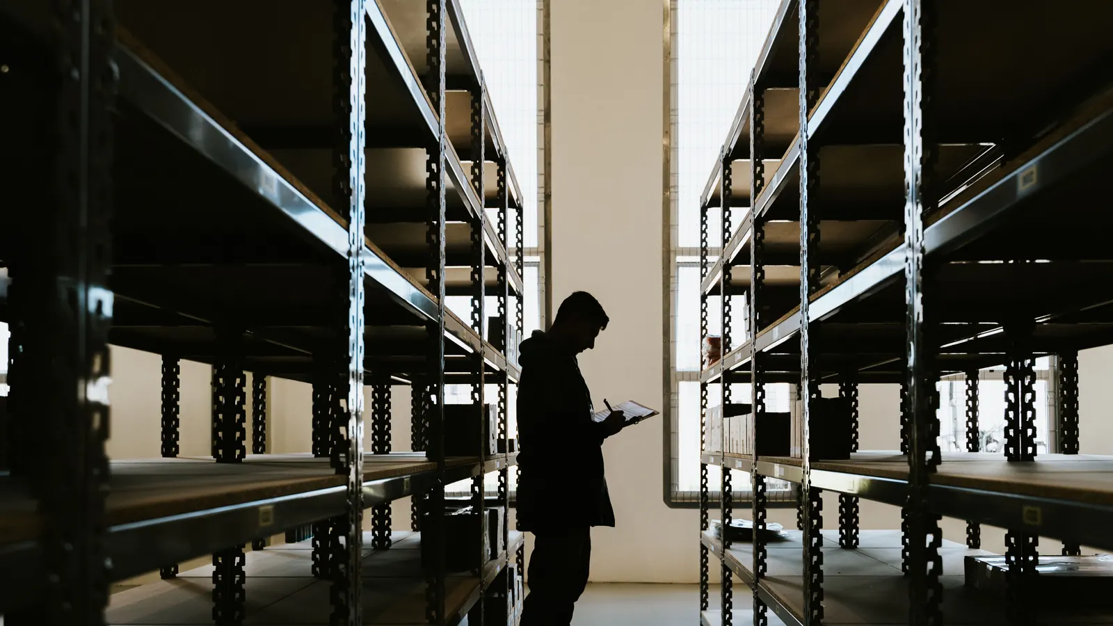 A silhouetted worker checking a tablet between rows of empty warehouse shelving