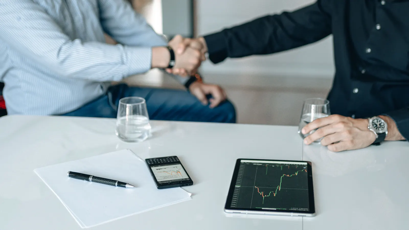 Two people shaking hands across a meeting table set with documents and a tablet