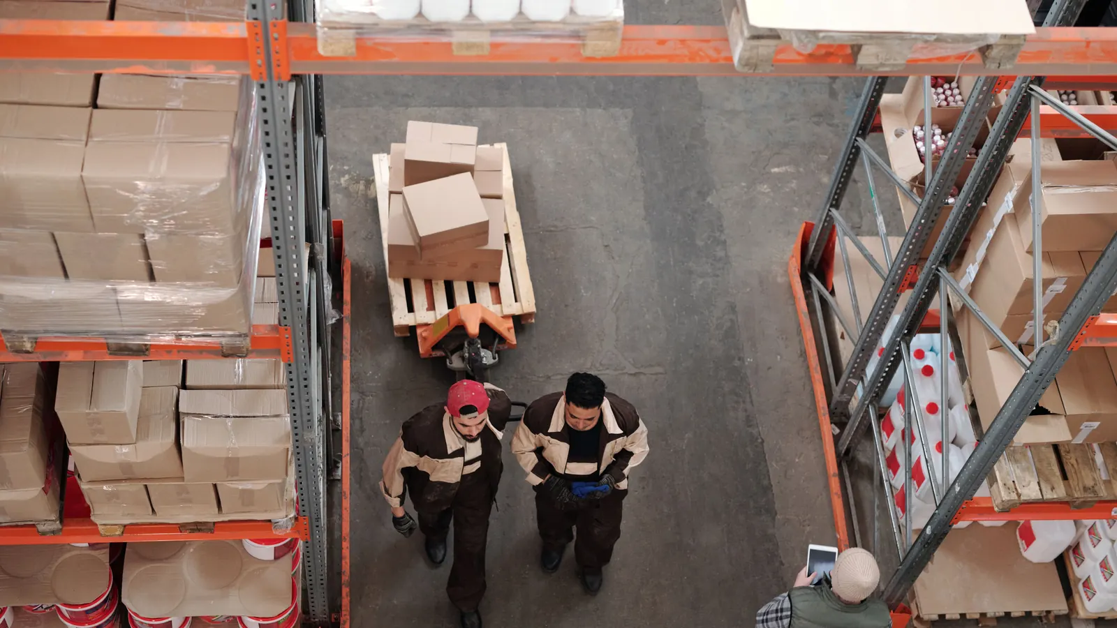 Overhead view of two warehouse workers pulling a pallet of boxes between stocked shelves