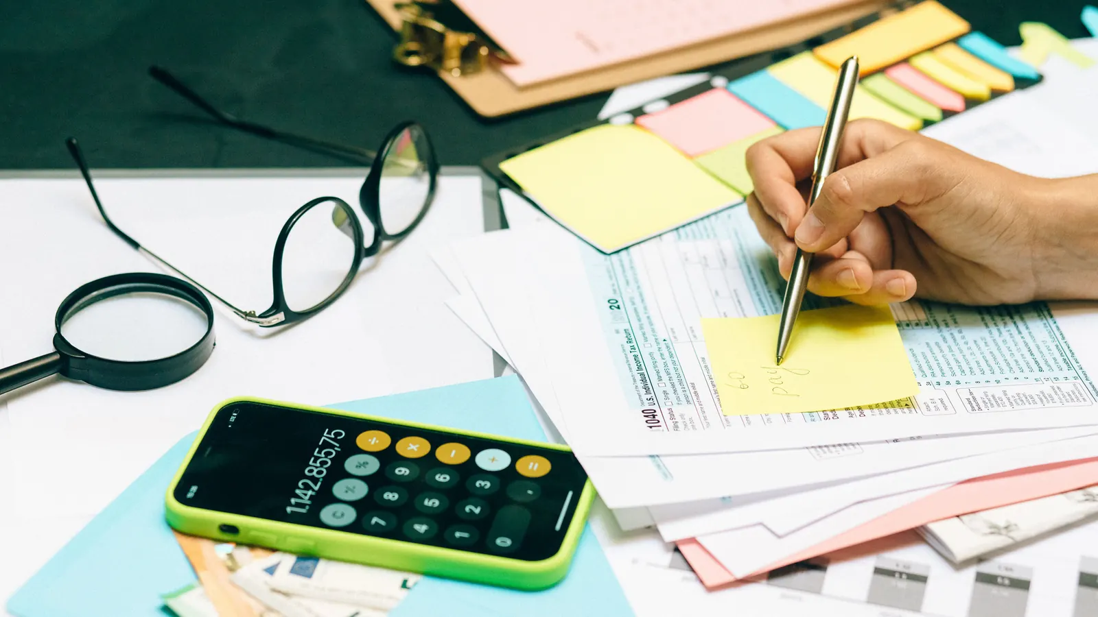 A hand annotating tax return paperwork with a pen, beside a calculator, reading glasses and a magnifying glass