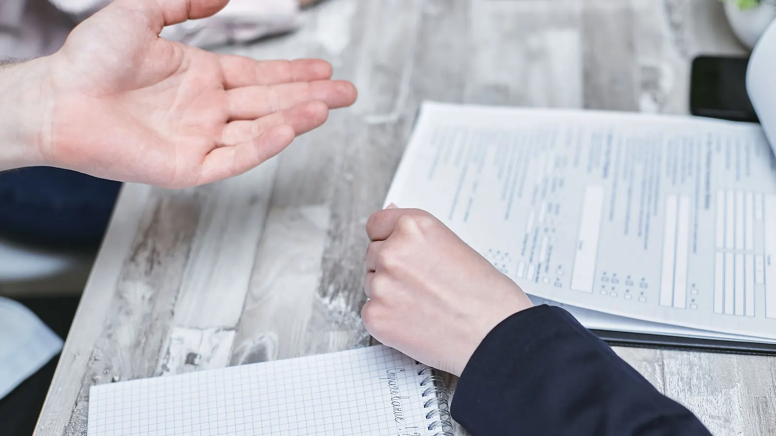 Two people at a desk exchanging a handshake gesture over paperwork and a notebook