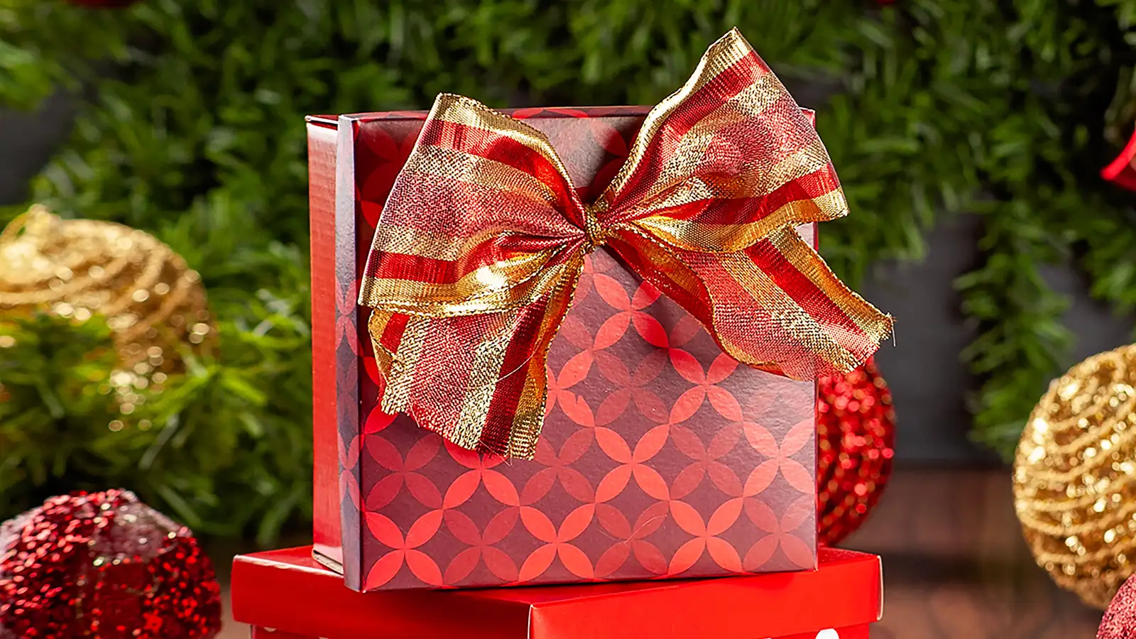 A red Christmas gift box tied with a gold ribbon among baubles and pine branches