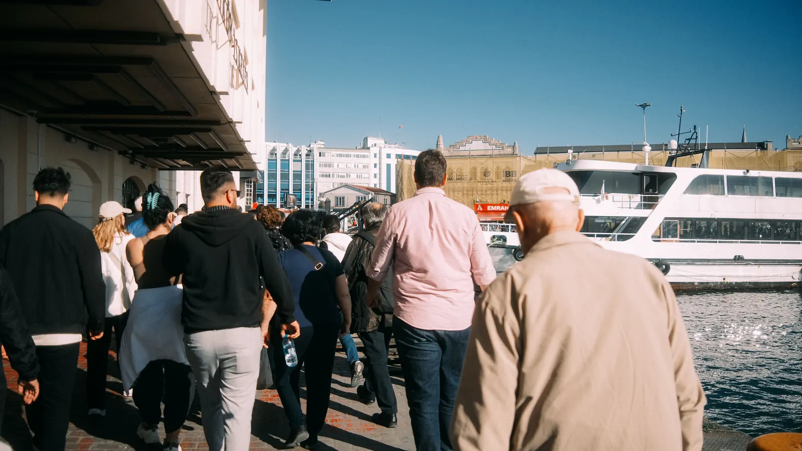 A group of people walking along a waterfront promenade past a docked ferry