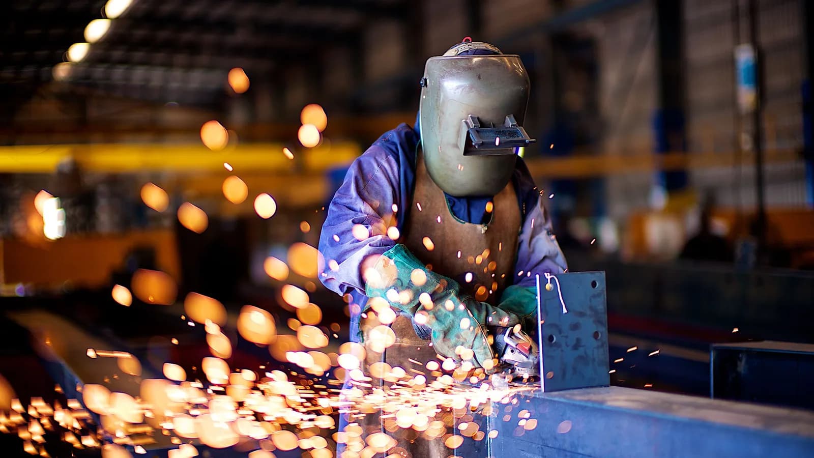 Factory worker in a welding mask grinding steel, with sparks flying across the workshop